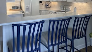 A modern kitchen with white cabinets, a central island with bar stools, a microwave, and a refrigerator. - CottageVacations4u "Beach Be With You"- ocean view- pool club-200 FT to beach! (Ocean Isle Beach)