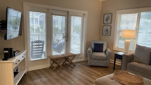 A living room with a sofa, armchair, and ottoman, a television, and a window with blinds. - CottageVacations4u "Beach Be With You"- ocean view- pool club-200 FT to beach! (Ocean Isle Beach)