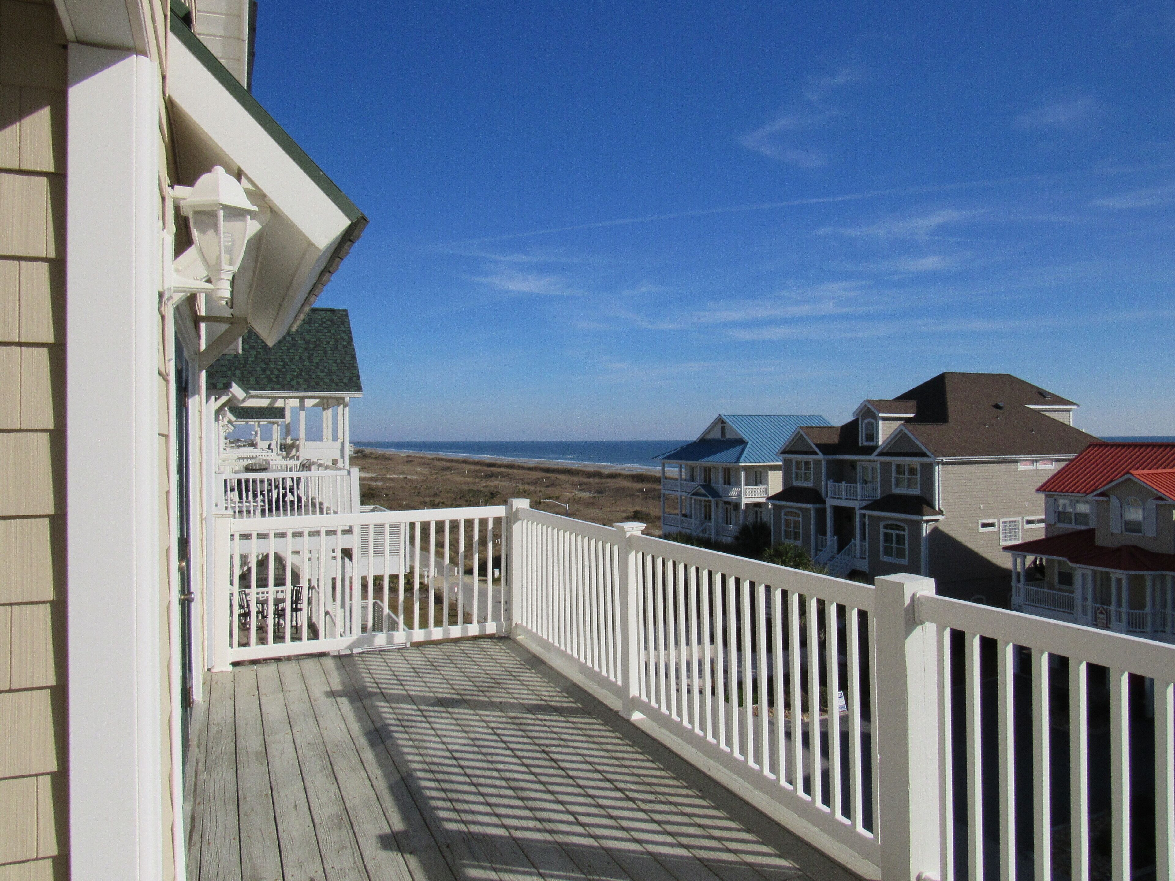 A wooden deck with a white railing, overlooking a beach and houses.
