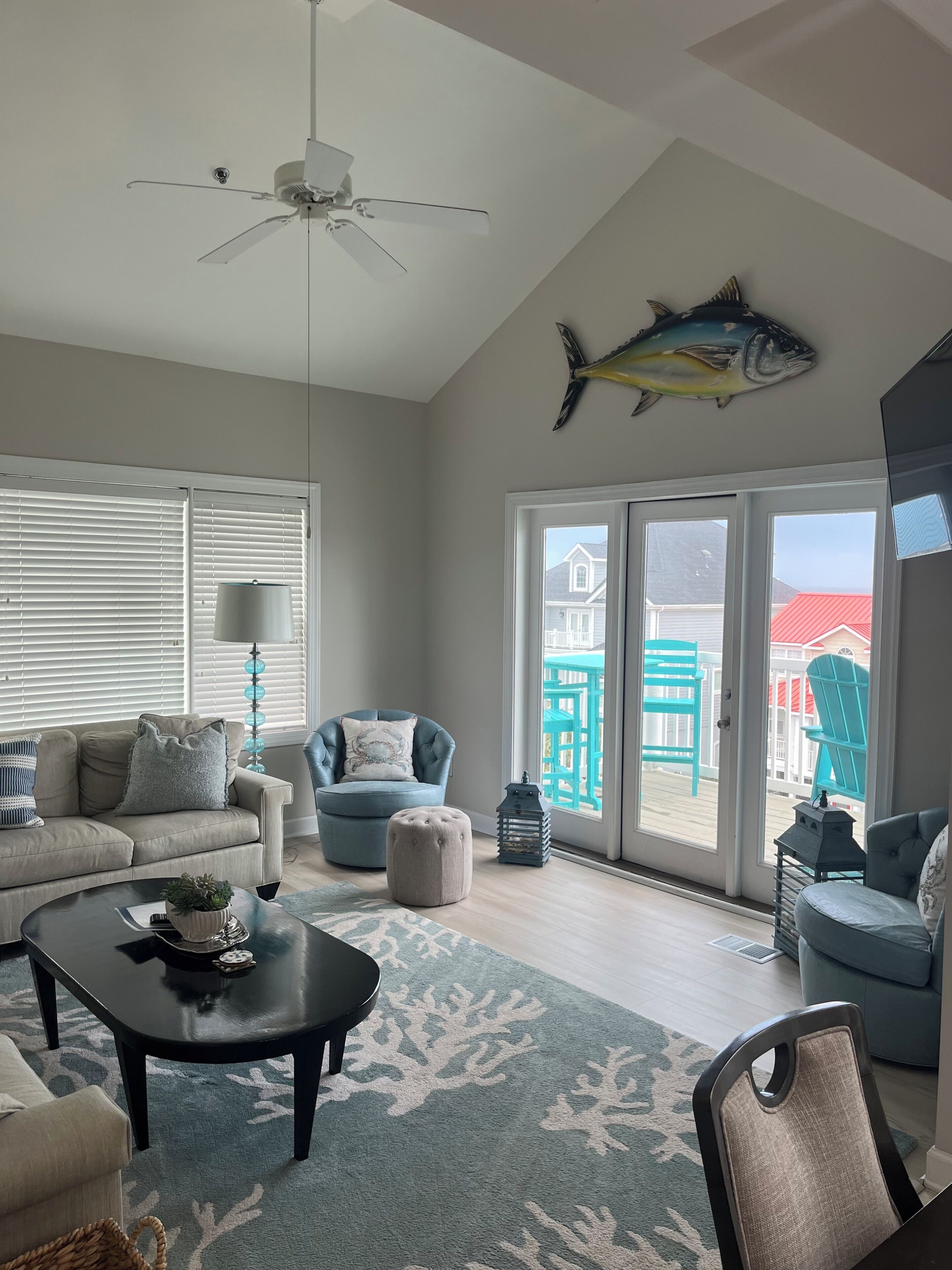 A living room with a ceiling fan, a fish decoration, a blue armchair, a sofa, and a view of the outside deck.