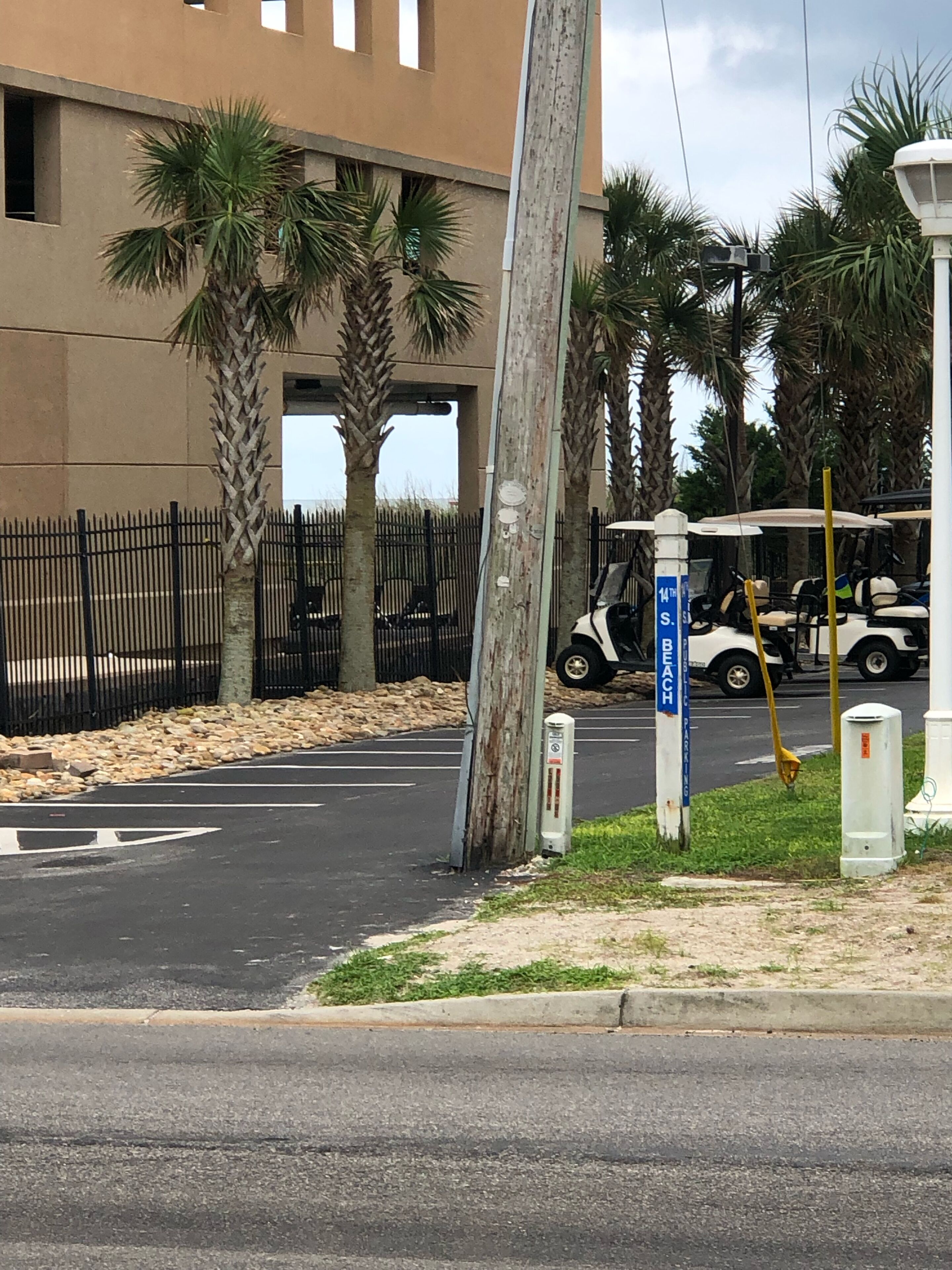A street with a building, palm trees, a golf cart, and a sign indicating a beach parking area.