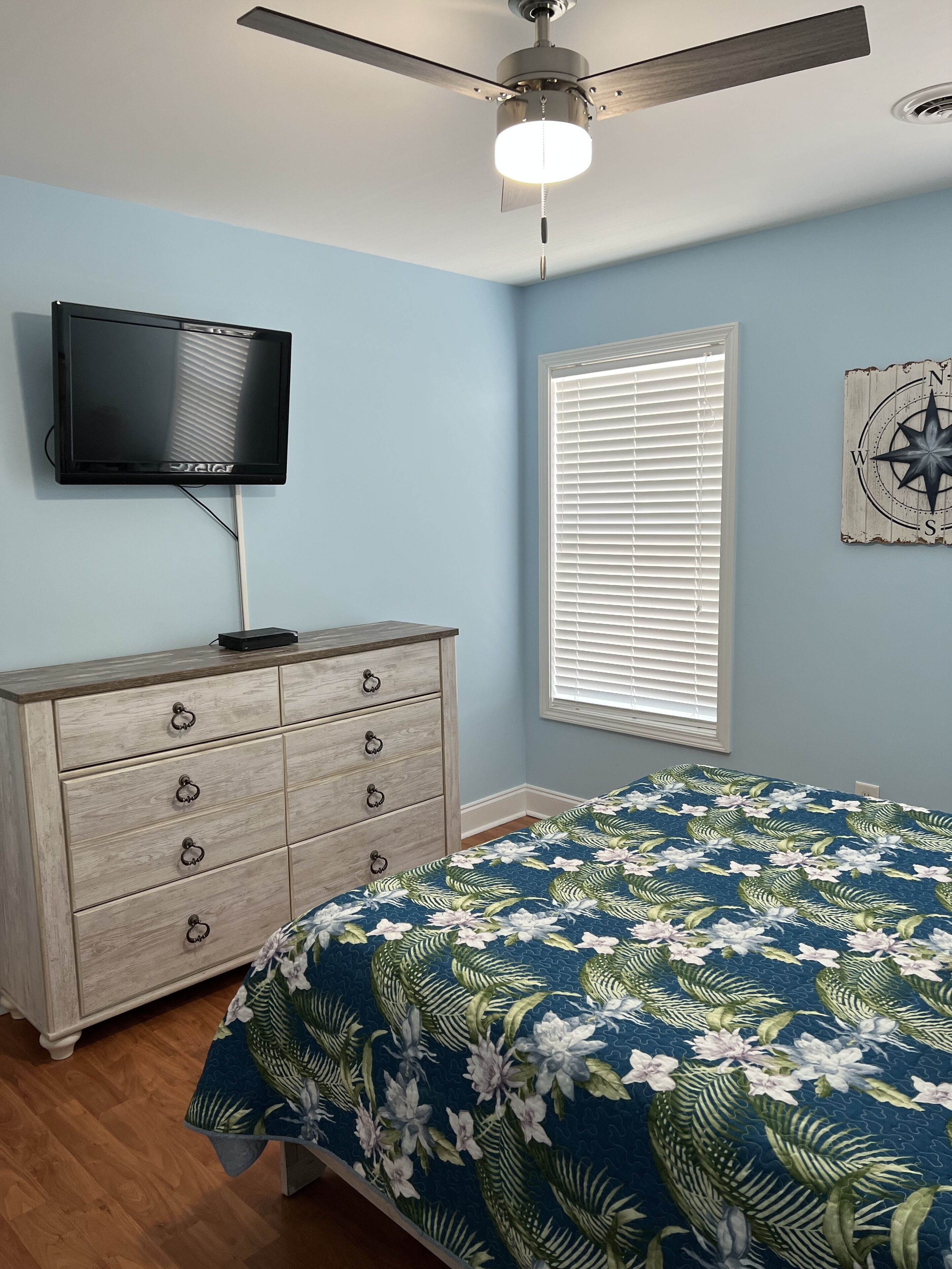 A bedroom with a bed, a dresser, a ceiling fan, a television, a window with blinds, and a wall decoration.