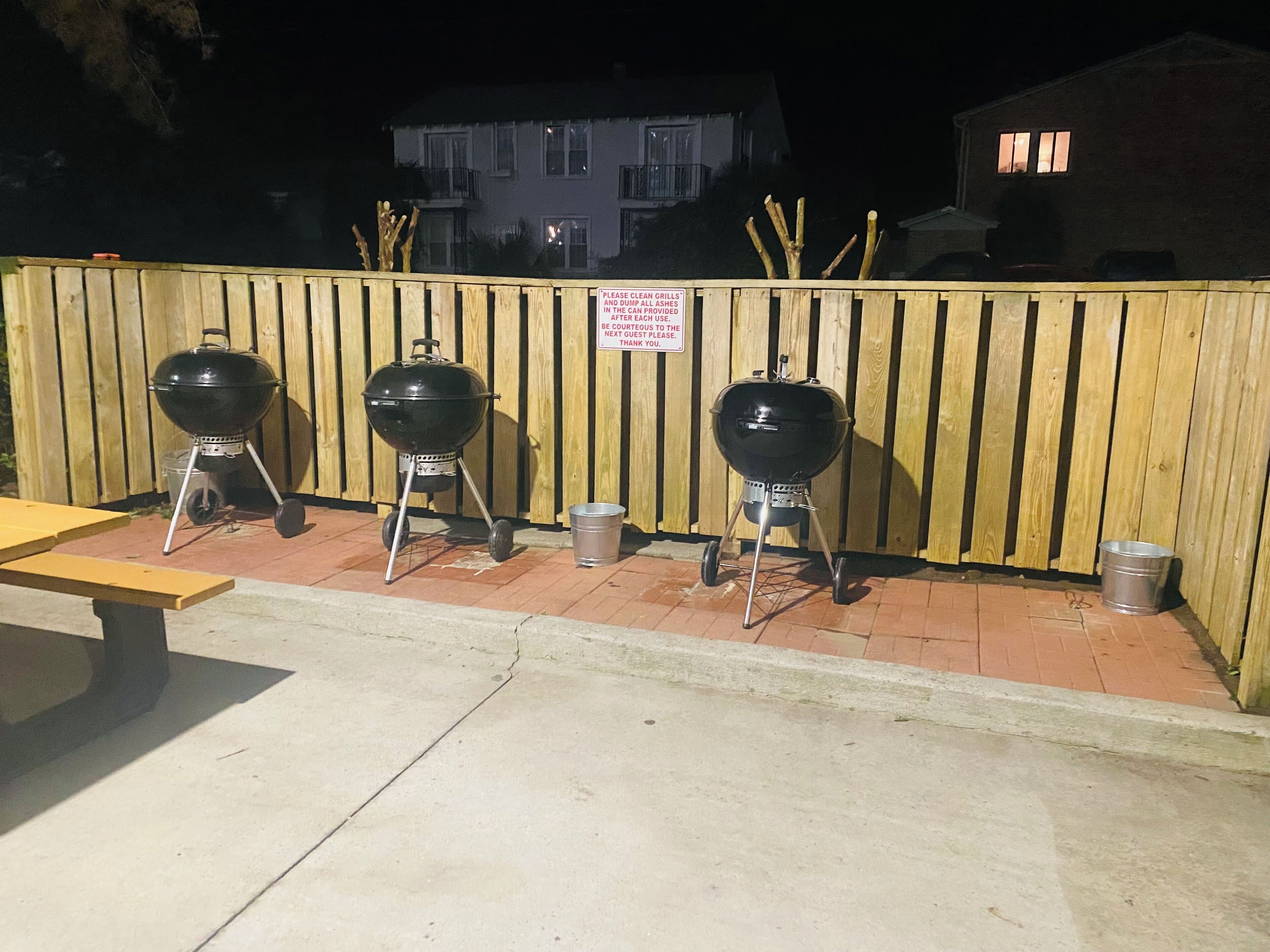 Three portable grills and a picnic table against a wooden fence.