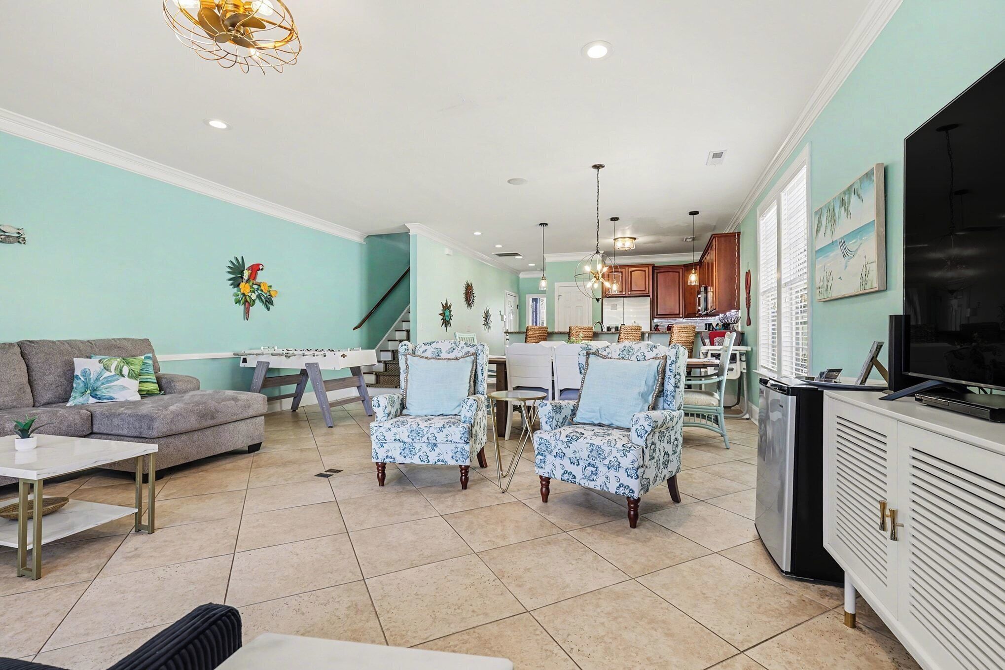 A spacious living room with a sofa, two armchairs, a coffee table, and a TV. A kitchen is visible in the background with a dining table and chairs.