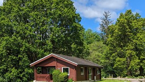 A small, single-story cabin with a porch, surrounded by lush green trees and a clear blue sky. - Stylish, Comfy & Cosy log cabin in National Park, dogs & families welcome! (Rashfield (near Dunoon))