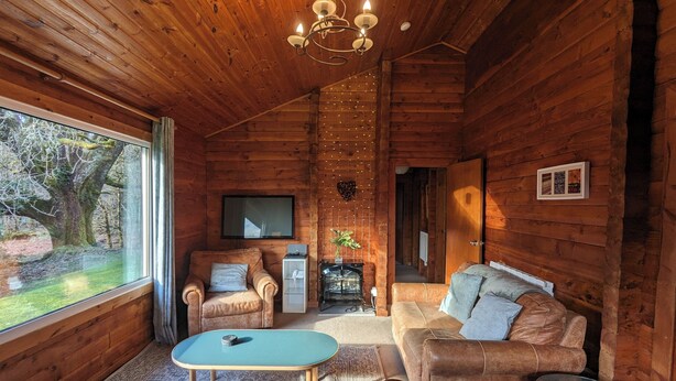 A cozy log cabin interior with a wooden ceiling, a chandelier, a fireplace, and a large window overlooking a tree. - Stylish, Comfy & Cosy log cabin in National Park, dogs & families welcome! (Rashfield (near Dunoon))