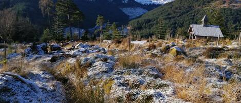 A mountainous landscape with a small cabin surrounded by snow-covered terrain and evergreen trees.