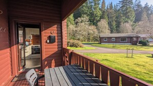 A wooden cabin with a porch, a dining table, and a view of a forest and another cabin in the distance. - Stylish, Comfy & Cosy log cabin in National Park, dogs & families welcome! (Rashfield (near Dunoon))