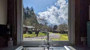 A kitchen with a large window offering a view of a residential area with houses and trees. - Stylish, Comfy & Cosy log cabin in National Park, dogs & families welcome! (Rashfield (near Dunoon))