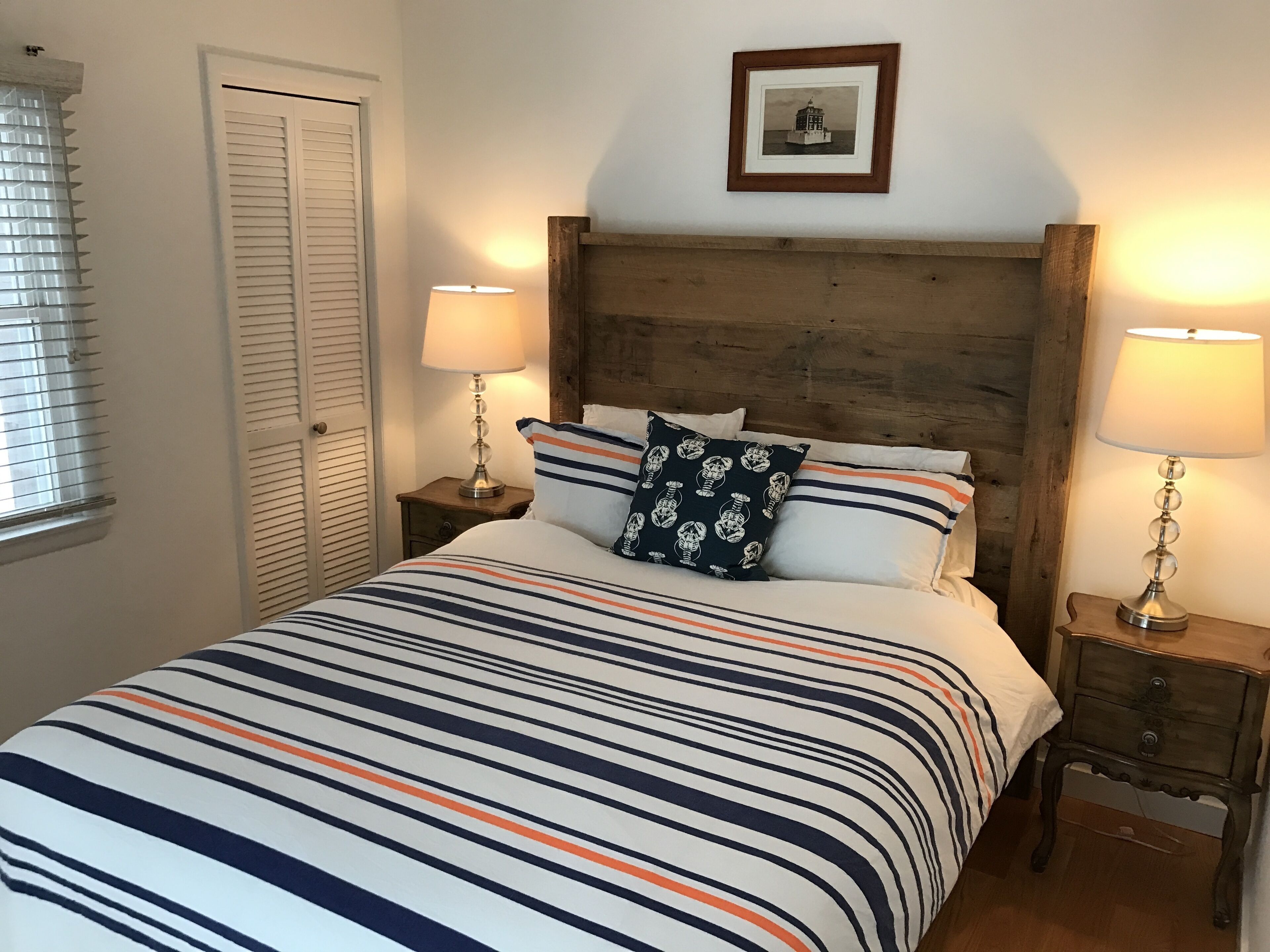 A bed with striped bedding, two bedside lamps, a wooden headboard, a framed picture on the wall, and a window with blinds.