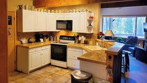 Cocina con armarios blancos, microondas, estufa y lavabo. Hay una ventana con vista a árboles y una silla en la sala.