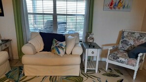 A living room with a beige sofa, a patterned armchair, a small side table, and a large tropical leaf-patterned rug. - Nader Vacation Rental (Little Torch Key)