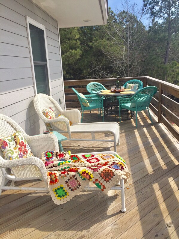 A wooden deck with wicker chairs and a table, a colorful crocheted blanket, and a view of trees.