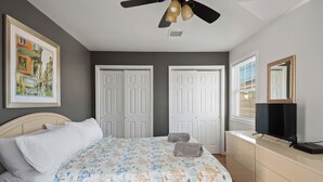 Un dormitorio con una cama, ventilador de techo, dos puertas blancas, cómoda, televisor y un cuadro en la pared.