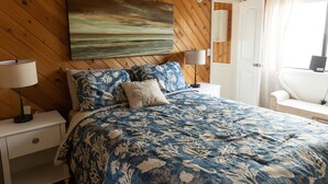 Une chambre à coucher avec un plafond lambrissé en bois, un grand lit recouvert d’une literie à motifs floraux, deux lampes de chevet, une table de chevet et une vue sur l’extérieur par une fenêtre ornée de rideaux.