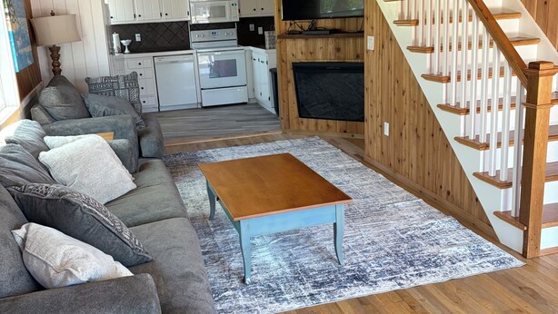 Un salon avec un canapé, une table basse et un téléviseur. Il y a une cuisine équipée et une cheminée. On peut voir un escalier en bois.