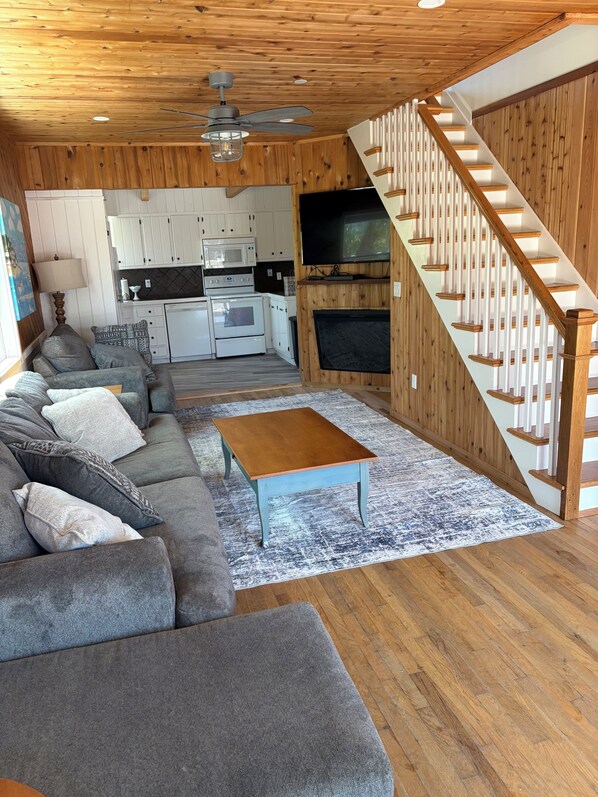 A living room with a sofa, coffee table, and TV. There's a kitchen with appliances and a fireplace. A wooden staircase is visible.