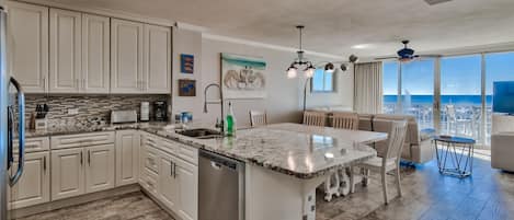 A modern kitchen with white cabinets, a granite countertop, and a built-in dishwasher. There is a dining area with a table and chairs, and a large window offering a view of the city.