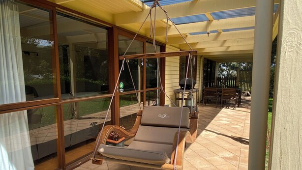 A covered patio with a swing chair, large windows, and a wooden table. - The Shelter bnb @ Robe SA (Robe)