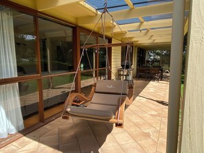 A covered patio with a swing chair, large windows, and a wooden table. - The Shelter bnb @ Robe SA (Robe)