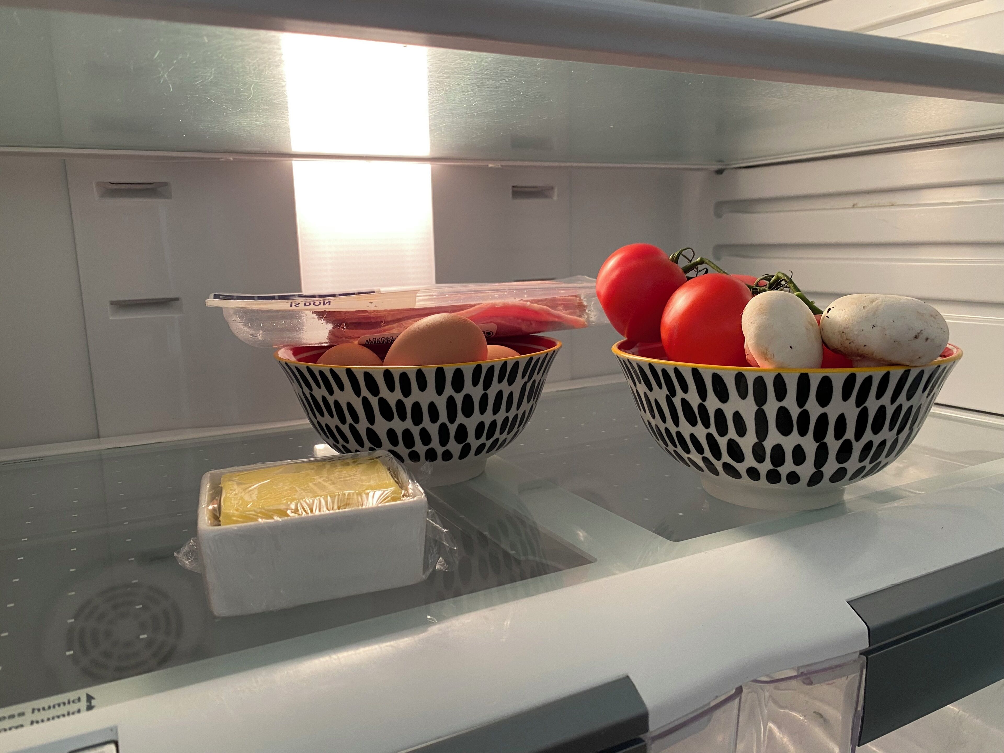 Twee versierde schalen op een koelkastplank, één met eieren en de ander met tomaten en champignons.