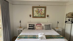 A neatly made bed with a striped blanket, a floral pillow, and a folded towel. There are two bedside tables with lamps, a wall-mounted picture, and a clothing rack. - The Shelter bnb @ Robe SA (Robe)