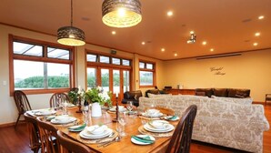 Un comedor con una mesa de madera preparada para una comida, ventanas amplias y un sofá al fondo.