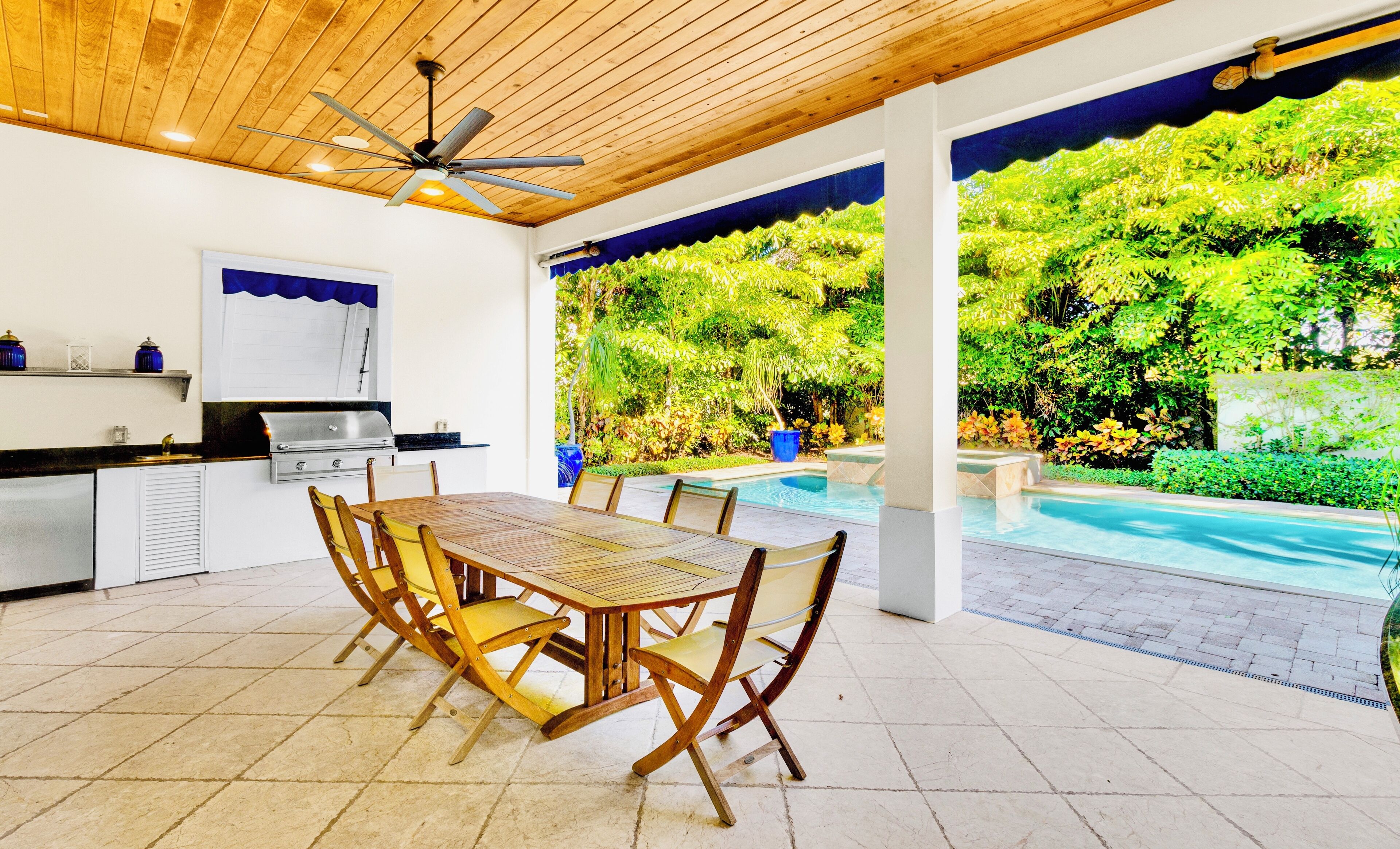 A covered patio with a pool, wooden furniture, and a grill area.