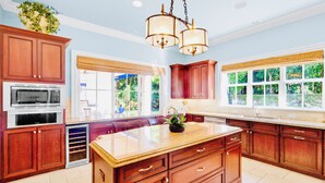 A spacious kitchen with wooden cabinets, a central island, and large windows offering a view of greenery. - Luxury Beach Retreat - 200 Steps to Beach - Heated Pool, Spa & Elevator (Jupiter)
