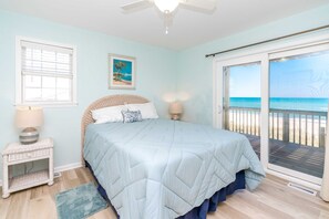 A bedroom with a bed, a nightstand, a lamp, a picture on the wall, and a view of the beach through a sliding door.