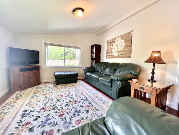 Una sala de estar con un sofá de cuero, un televisor de pantalla plana, un mueble de madera, un pequeño reposapiés, un tapete con motivos florales, una lámpara de pared y una estantería.
