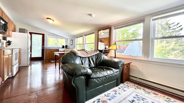 A spacious living room with a leather couch, wooden flooring, a dining area, and large windows offering a view of the outdoors. - Quiet oasis across the street from Acadia and just steps from the harbor (Seal Harbor)