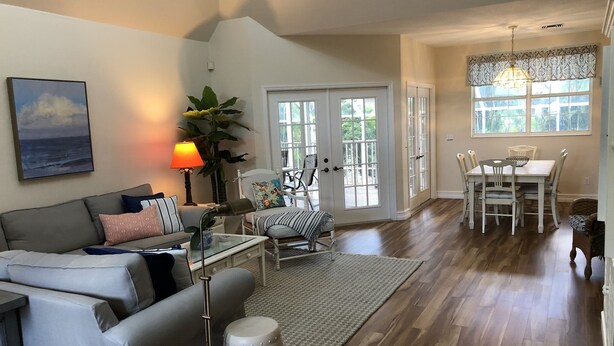 A spacious living room with a grey sofa, a coffee table, and a lamp. There is a dining area with a table and chairs, and a large window with curtains. - Key West Style Florida Charm w Heated Private Salt Pool, close walk to Beach (Marco Island)