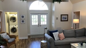 A living room with a grey sofa, a wicker chair, a glass coffee table, and a floor lamp. There is a door with a window, a washing machine, and a framed picture on the wall. - Key West Style Florida Charm w Heated Private Salt Pool, close walk to Beach (Marco Island)