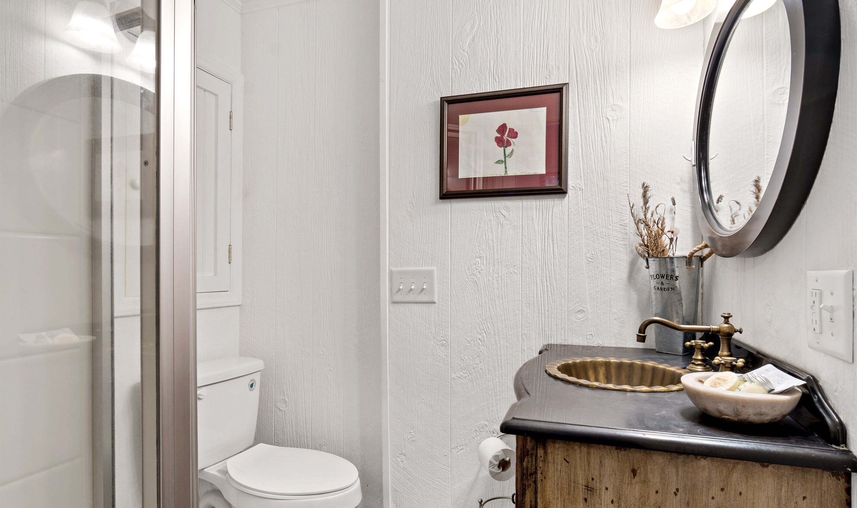 A bathroom with a white toilet, a wooden vanity, a mirror, and a framed picture of a flower.