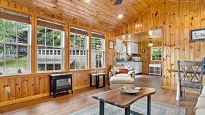 Un intérieur de chalet chaleureux avec de grandes fenêtres, une cheminée, un canapé et un coin cuisine.