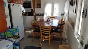 A compact kitchen with a microwave, sink, and white cabinets. A dining table with chairs is in the center, and a refrigerator is on the left. - The Poplars. 15 Steps & Your Toes Are In The Sand! (South Haven)