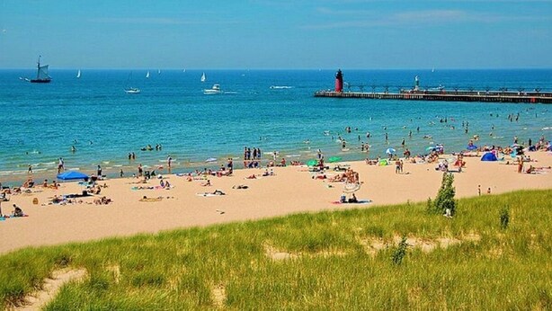 Een strand met mensen die zonnebaden en zwemmen, een steiger met een vuurtoren en zeilboten in de verte.