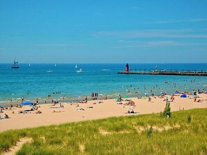 Una playa con gente tomando el sol y nadando, un muelle con un faro, y veleros a lo lejos.