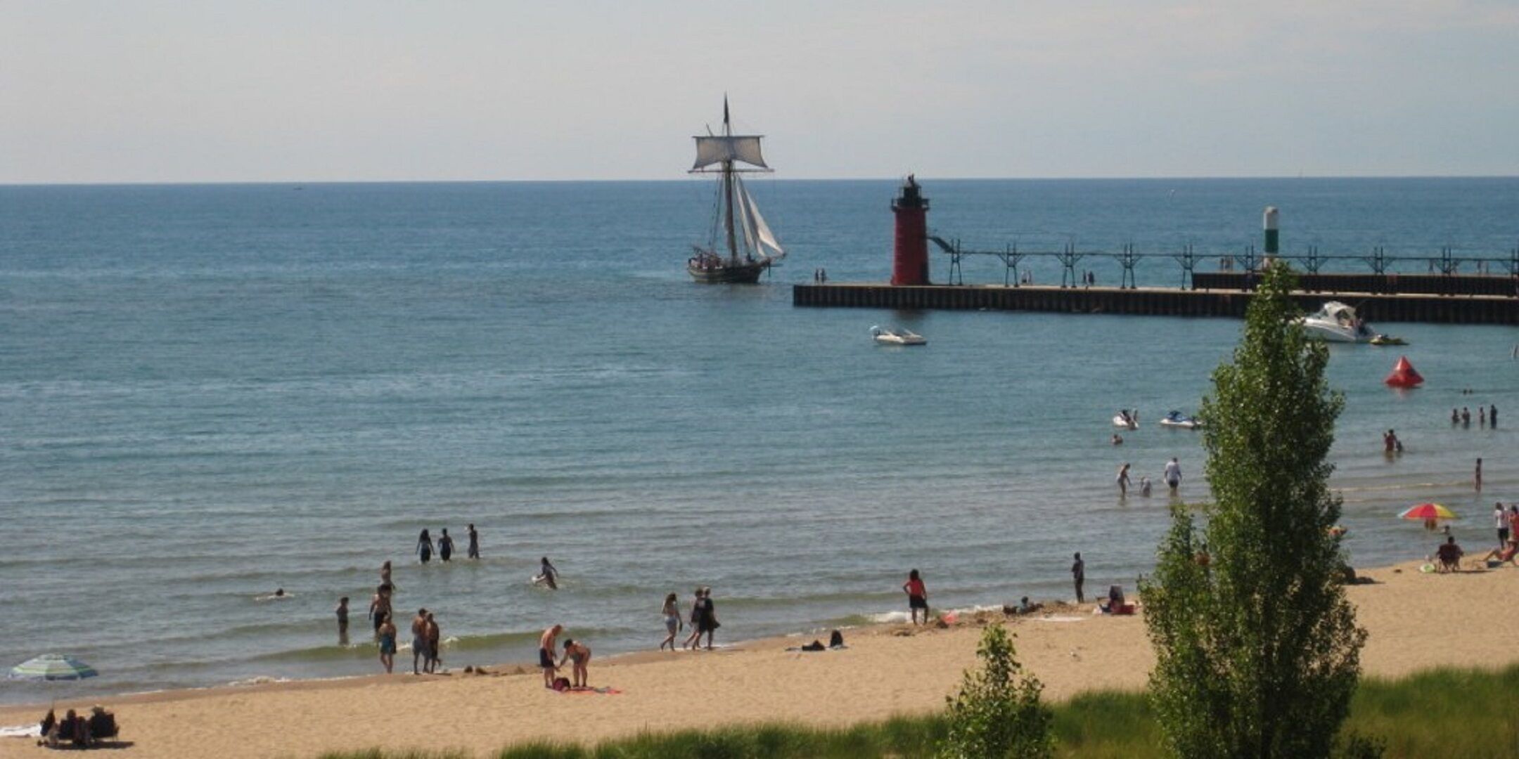 A beach with a lighthouse, a sailboat, and people enjoying the water.
