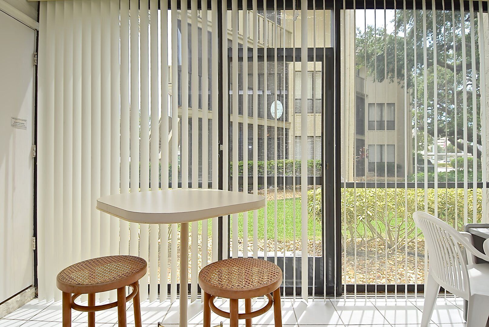 A balcony with two wicker chairs and a small table, a white door, and a view of a garden and buildings through a metal fence.