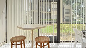 Un balcón con dos sillas de mimbre, una mesa pequeña, una puerta blanca y vistas a un jardín y edificios a través de una cerca de metal.