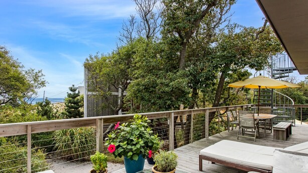 Eine Holzterrasse mit Gartenmöbeln, Blumentöpfen und Blick auf Bäume und Hügel.
