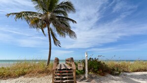 Un escalier en bois menant à une plage où se trouve un palmier et un panneau d’avertissement.