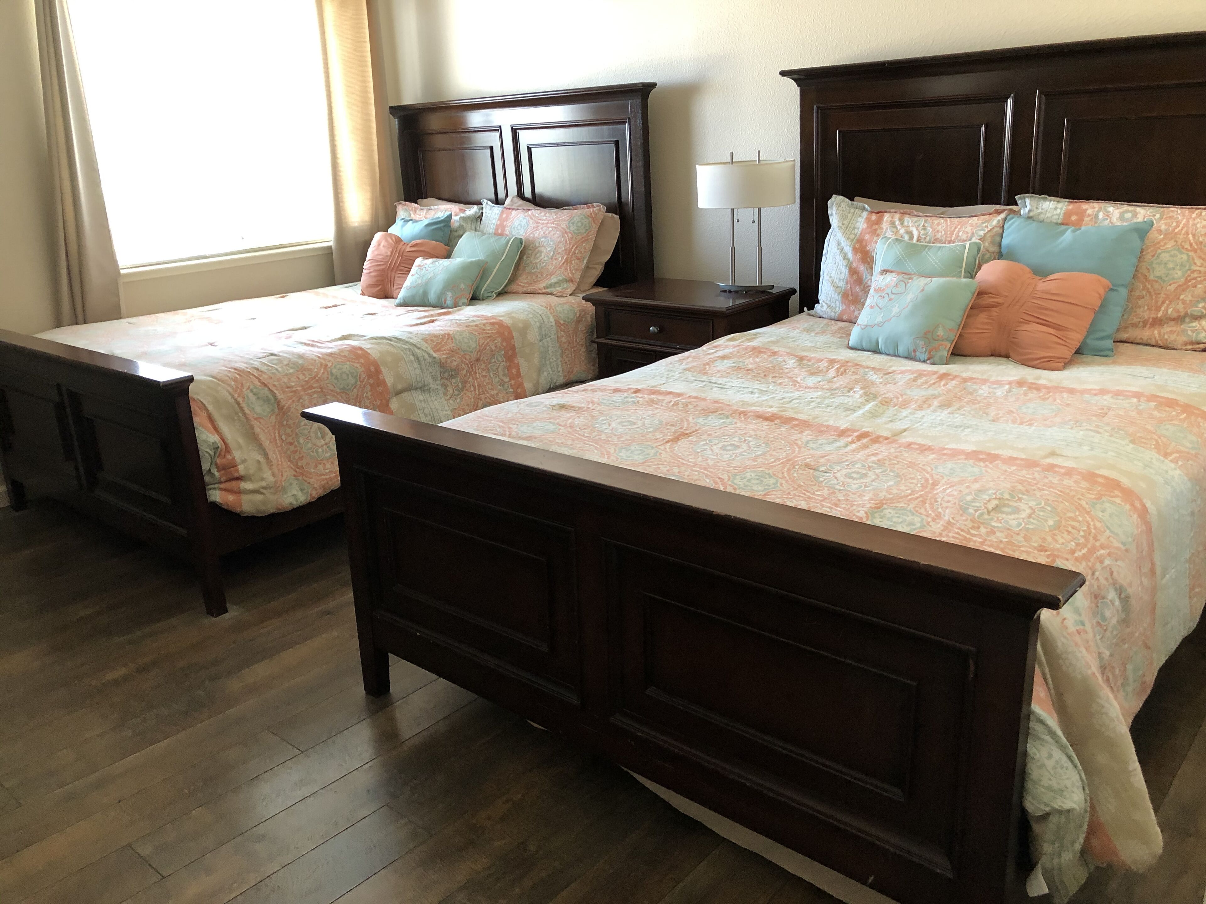 Une chambre d’hôtel avec deux lits en bois, chacun agrémenté de draps à motifs et de plusieurs oreillers. Une table de chevet avec une lampe se trouve entre les deux lits.