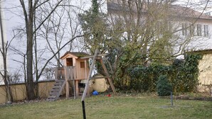 Una casita de juegos de madera con una escalera, rodeada de árboles y un patio cubierto de césped.