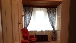 Un interior con piso de madera, un sillón rojo, un mueble de madera oscura y una ventana con cortinas estampadas.