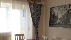 Un área de comedor con una mesa de madera y sillas blancas, un banco, cortinas estampadas y un cuadro de un paisaje urbano en la pared.