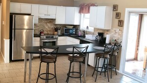 A kitchen with white cabinets, a stainless steel refrigerator, a microwave, and a coffee maker. There are three bar stools and a table. - Perfect Getaway In The Poconos! (House #1) HotTub !! Gameroom!! WiFi !! (Albrightsville)
