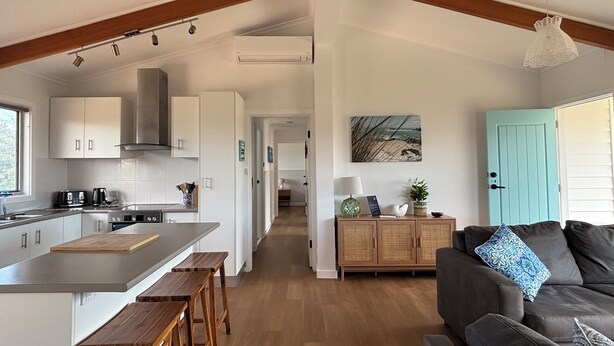 Une cuisine moderne avec des meubles blancs, un îlot central, des tabourets en bois et un coin repas agrémenté d’un canapé et de coussins aux couleurs foncées.
